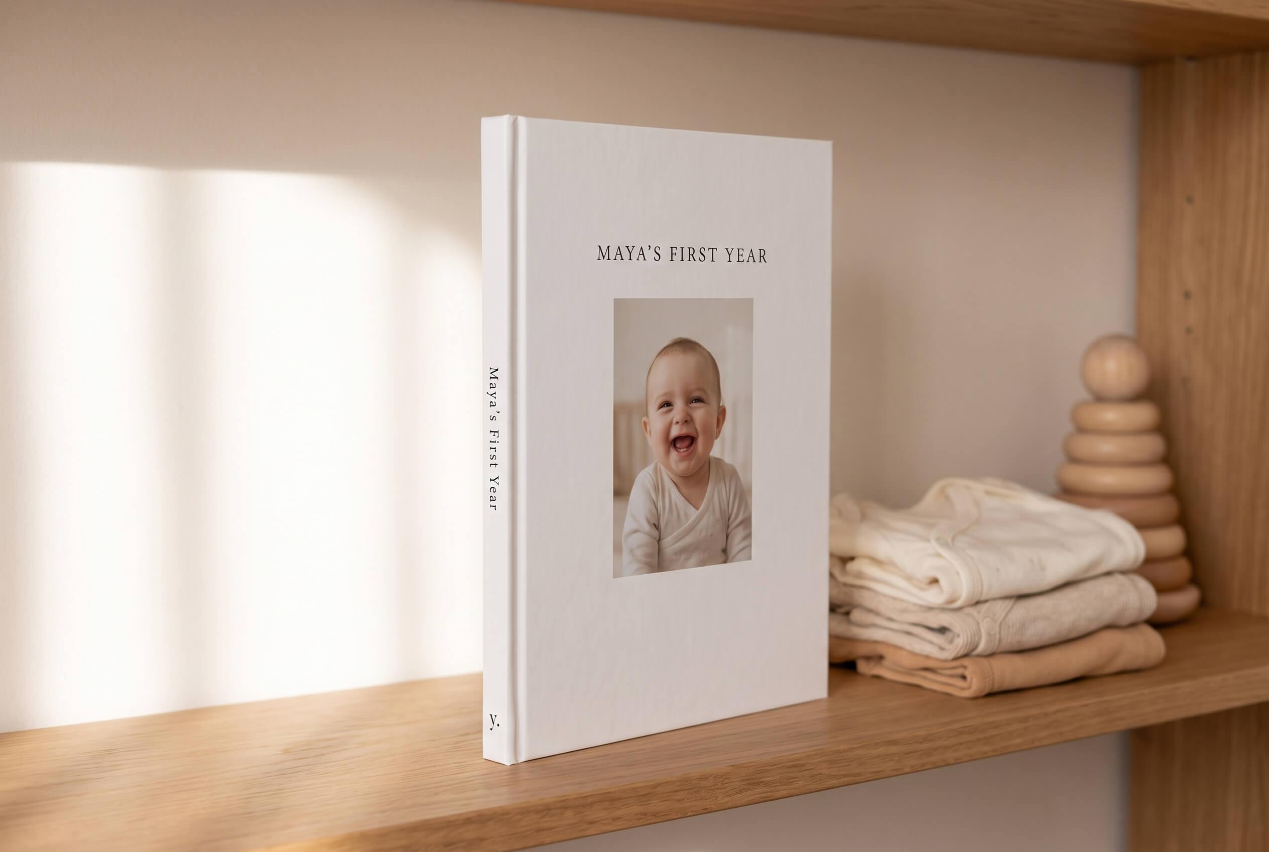 Closed yeerbook titled Maya's First Year standing on a nursery shelf beside folded baby clothes